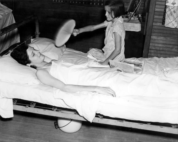 A little girl in 1938 fans her sick mother, who works in a box factory. Huey Long greatly expanded access by the rural poor to professional healthcare.